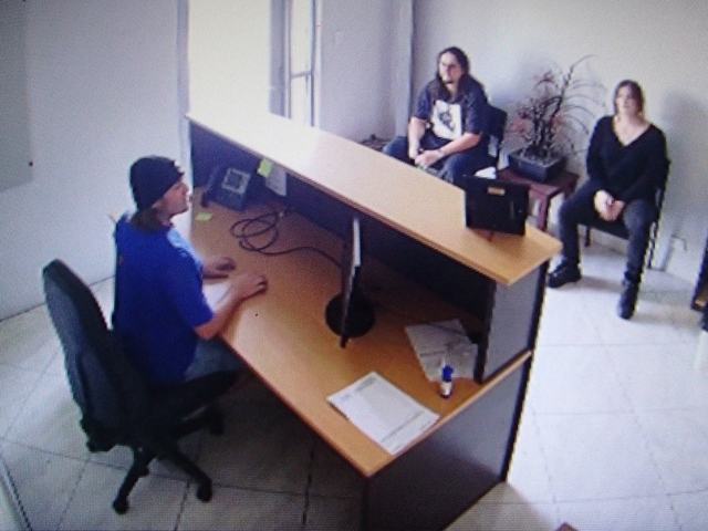 CCTV view of an office reception, one band member at the front desk and two waiting in chairs