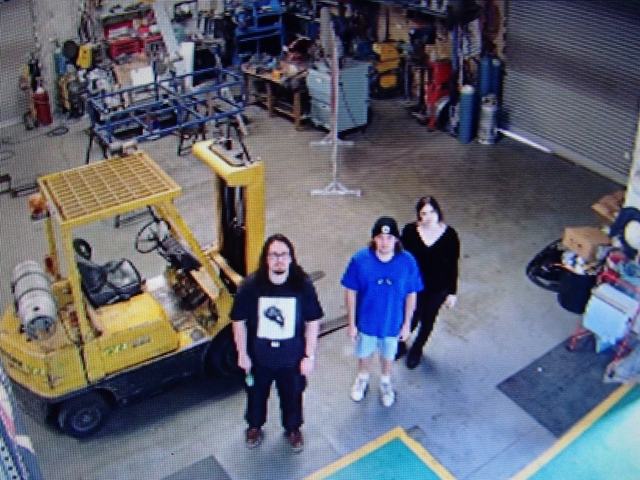 CCTV view of the three band members standing on a warehouse floor beside a yellow forklift